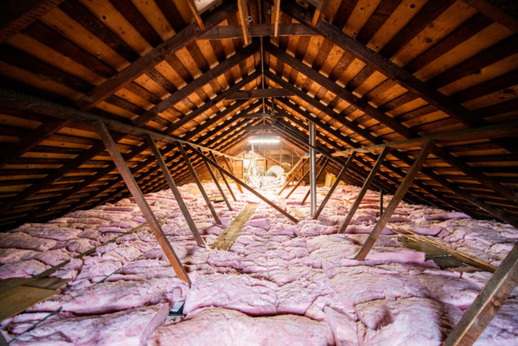 View of an attic with exposed wooden beams and soft pink insulation covering the floor, illuminated by a central light source.