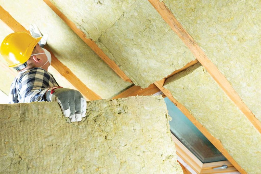 A man wearing a hard hat and helmet installs insulation in a construction setting, focused on his task.