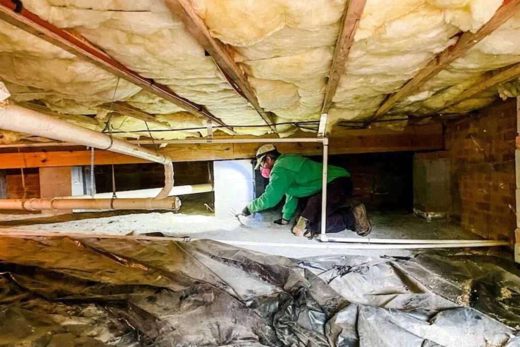 A worker in a green jacket inspects a crawl space, surrounded by insulation and pipes, with a flashlight illuminating the area.