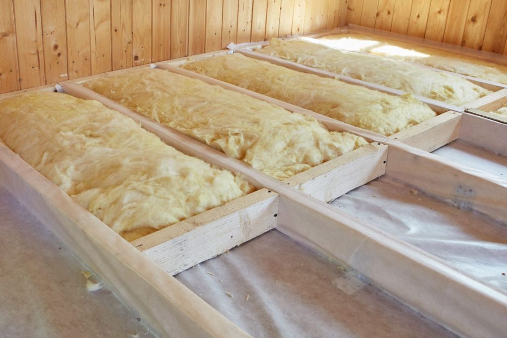 Insulation batts installed between wooden beams in a well-lit attic space, with wooden paneling in the background.