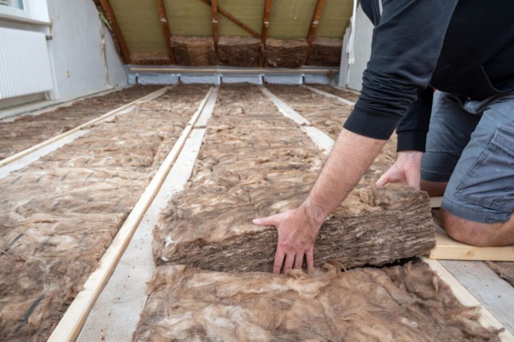 A man installs insulation material evenly on the attic floor between wooden beams.