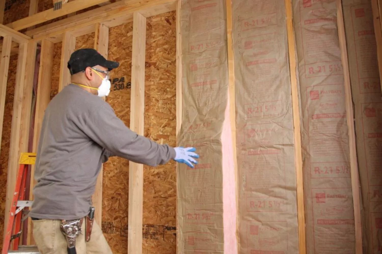 A worker wearing a mask and gloves installs insulation between wooden studs in a home under construction.