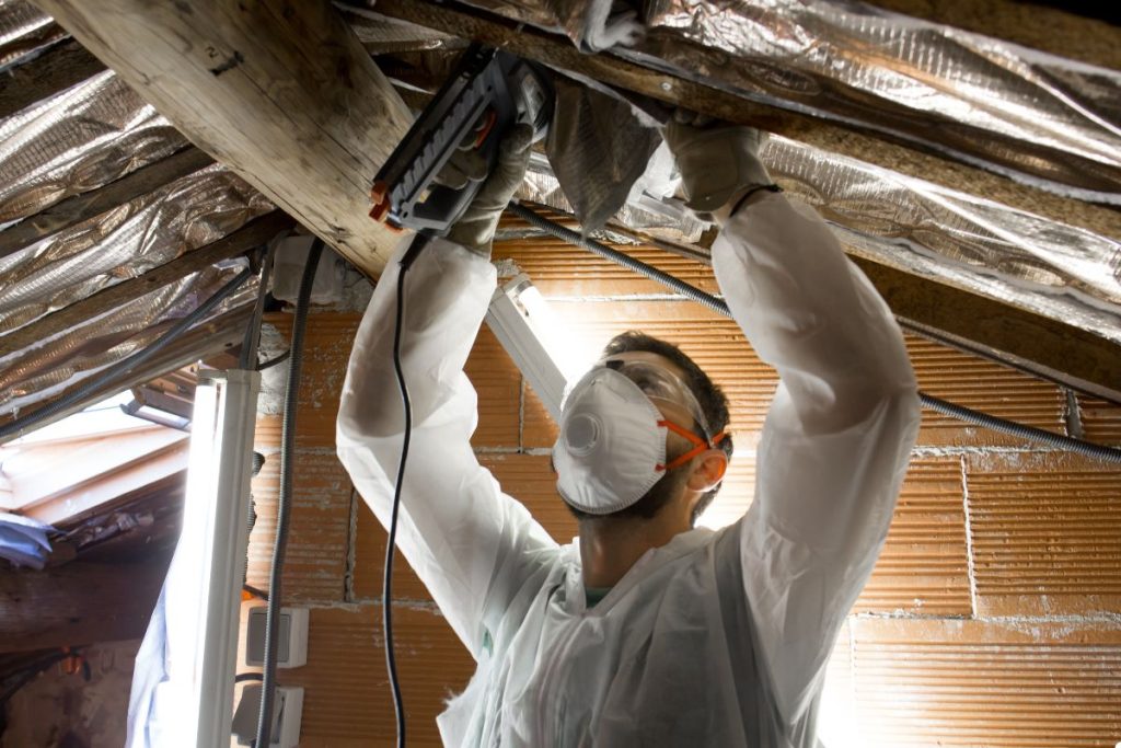 A man wearing a white coat and mask is engaged in work on a roof, utilizing various tools for maintenance tasks.