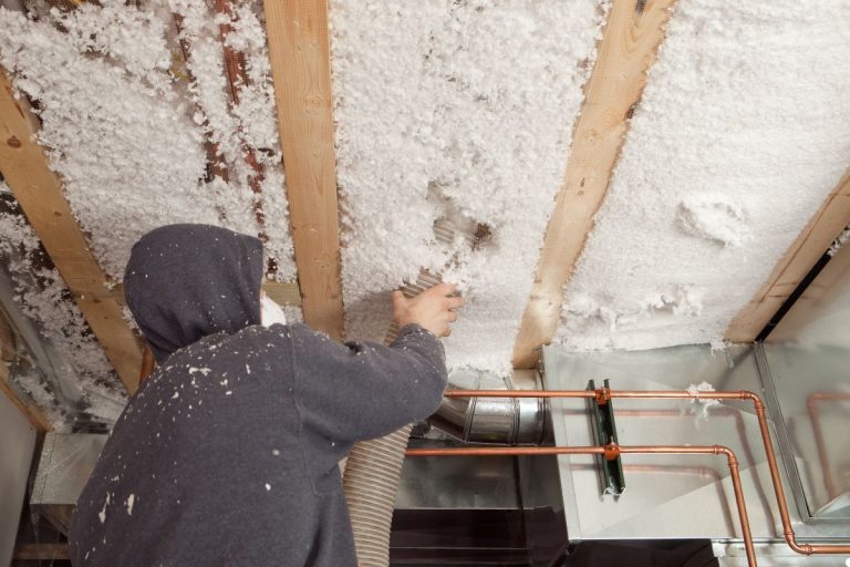 A man in a hoodie installs insulation on a ceiling, focused on his work with tools in hand.